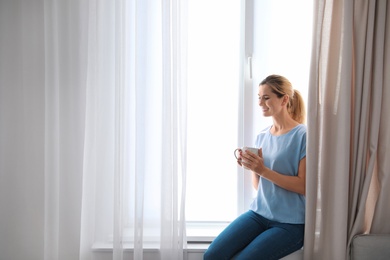 Woman holding cup of coffee near window with beautiful curtains at home. Space for text