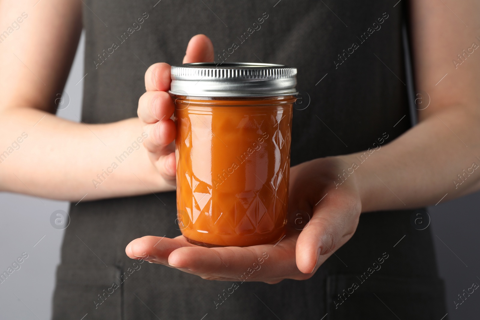 Photo of Woman holding glass jar of delicious persimmon jam on gray background, closeup