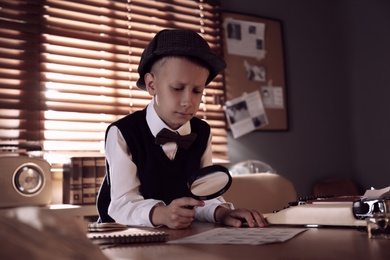Photo of Cute little detective exploring fingerprints with magnifying glass at table in office