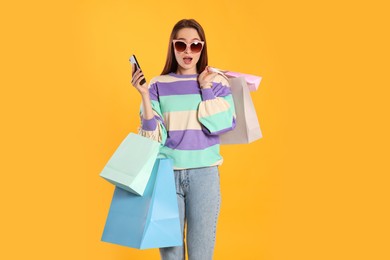 Photo of Surprised young woman with shopping bags and smartphone on yellow background. Big sale