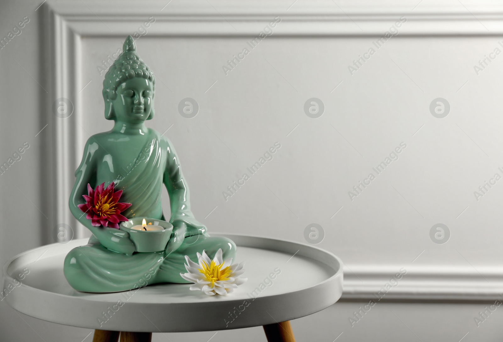 Photo of Buddha statue with burning candle and lotus flowers on white table. Space for text