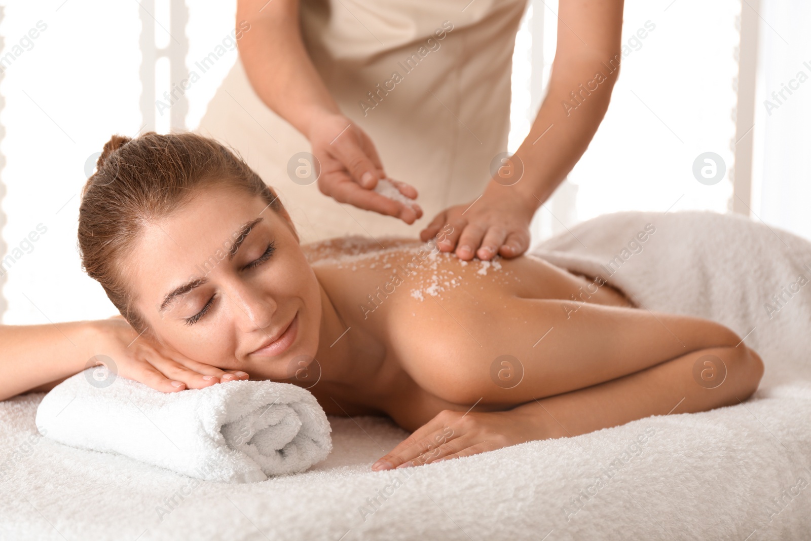 Photo of Young woman having body scrubbing procedure with sea salt in spa salon