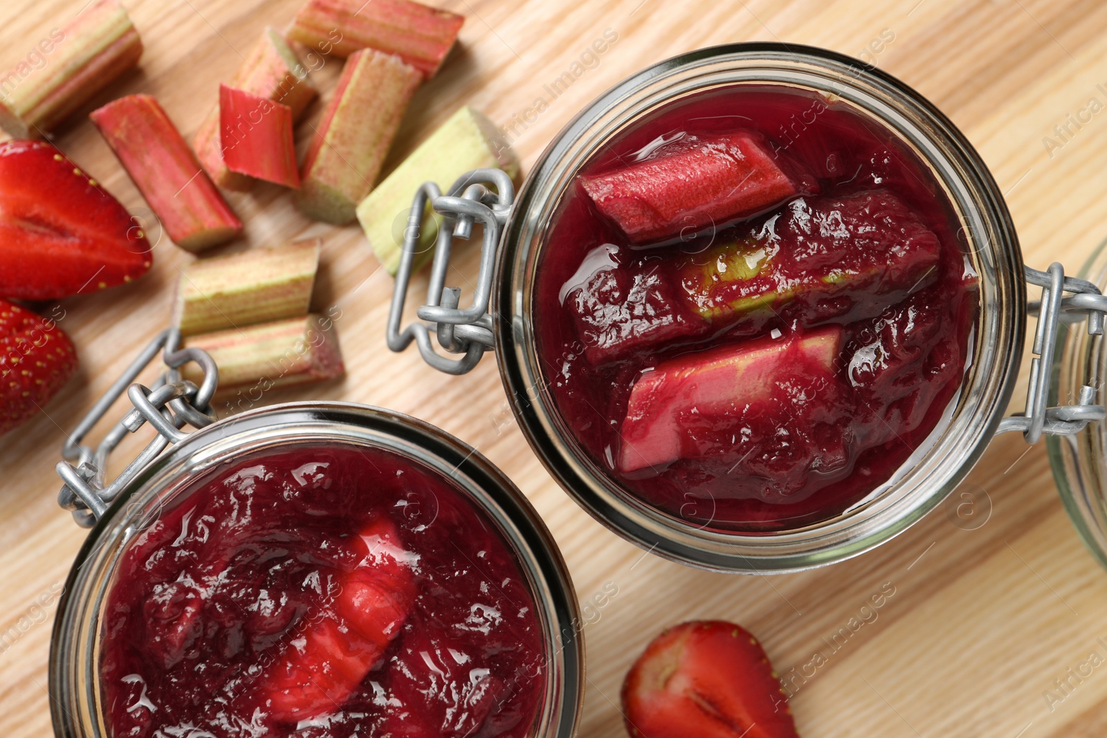 Photo of Jars of tasty rhubarb jam, cut stems and strawberries on wooden table, flat lay