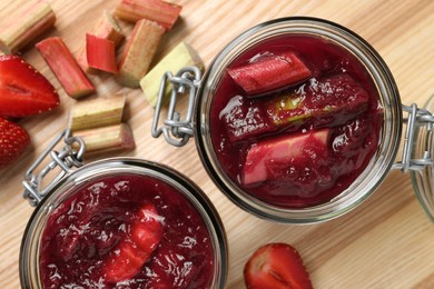 Jars of tasty rhubarb jam, cut stems and strawberries on wooden table, flat lay