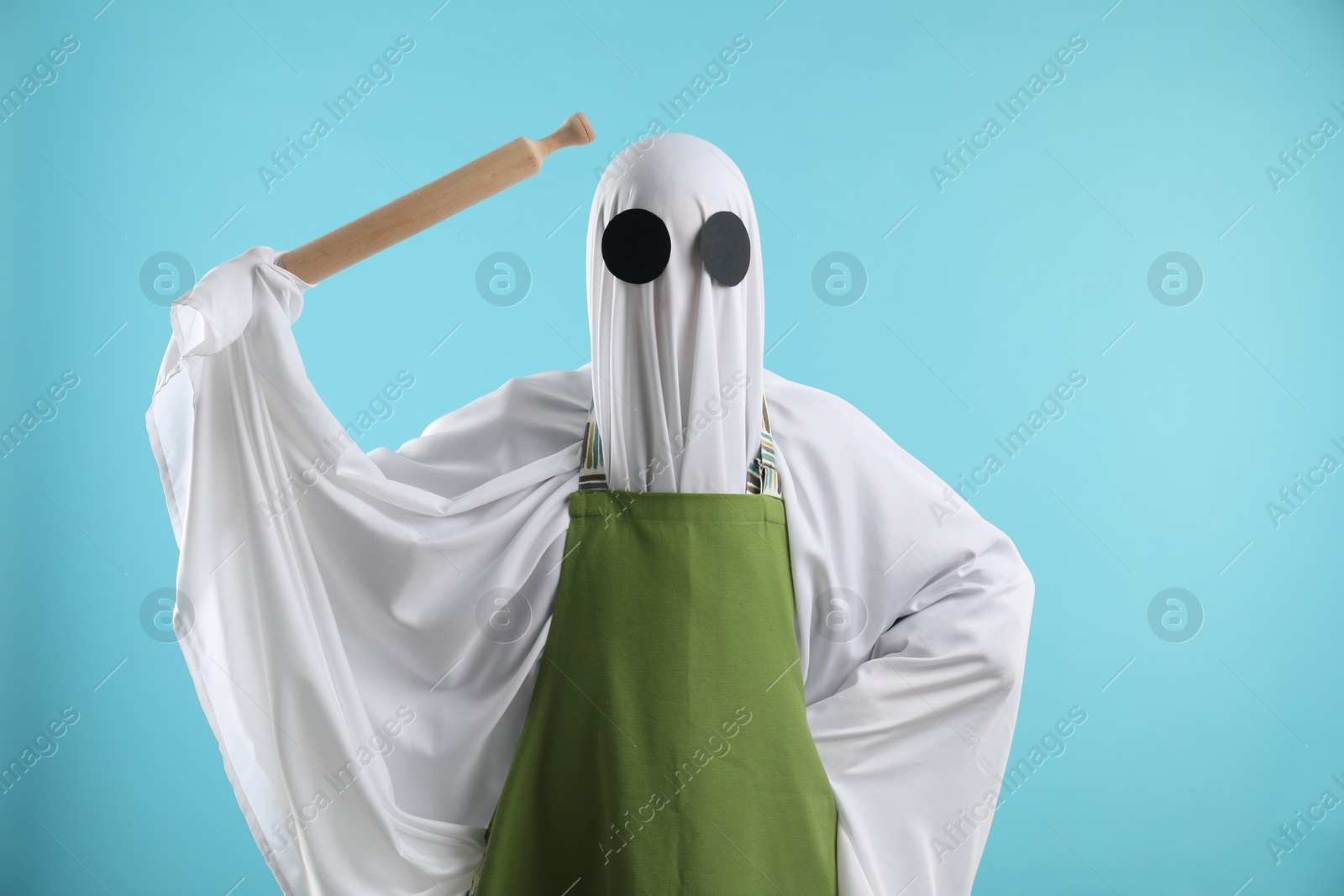 Photo of Woman in ghost costume and apron with rolling pin on light blue background