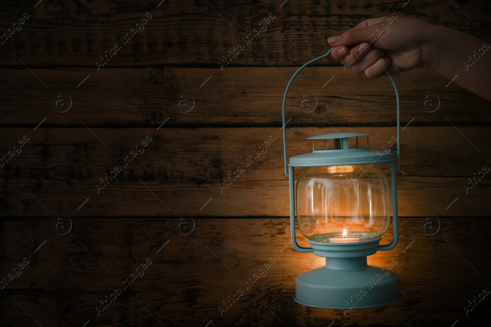 Photo of Woman holding lantern with burning candle against wooden wall in darkness, closeup. Space for text