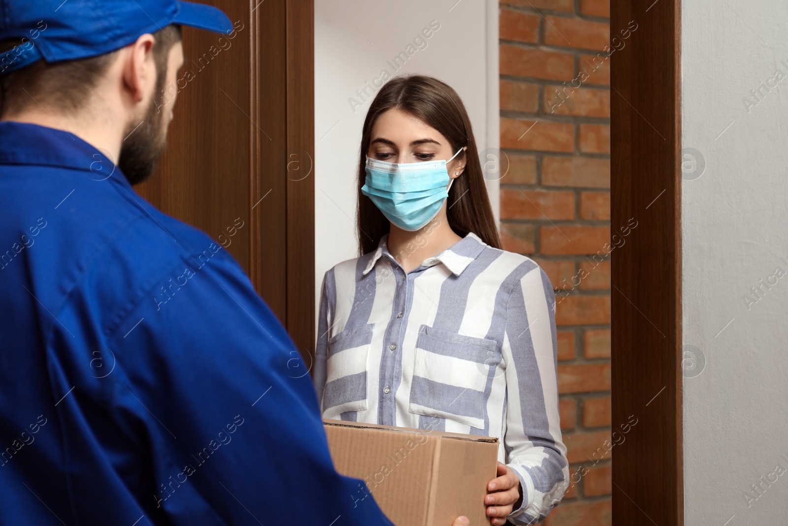 Photo of Young woman wearing medical mask receiving parcel from delivery man indoors. Prevention of virus spread