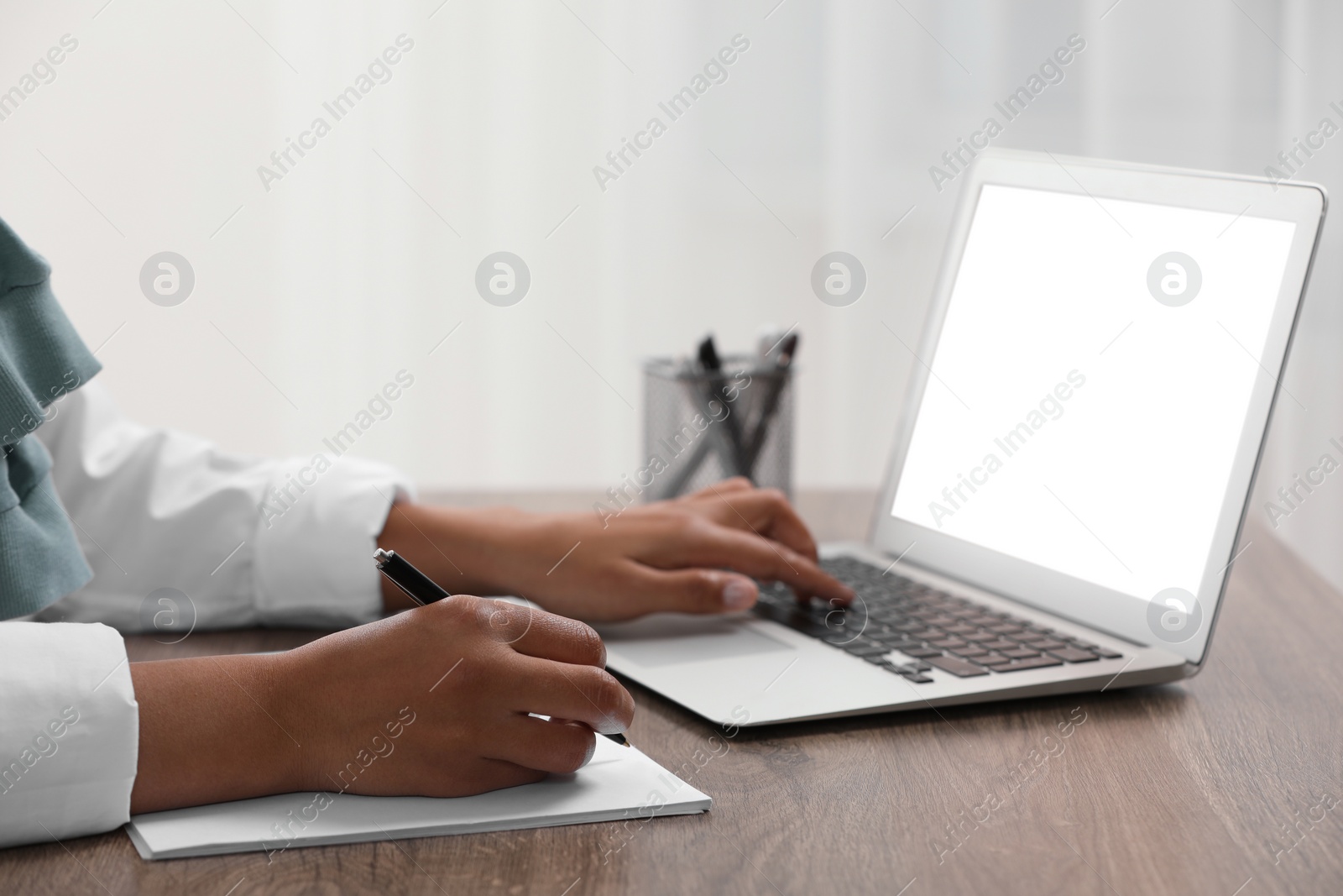 Photo of Woman writing notes while using laptop at wooden desk indoors, closeup