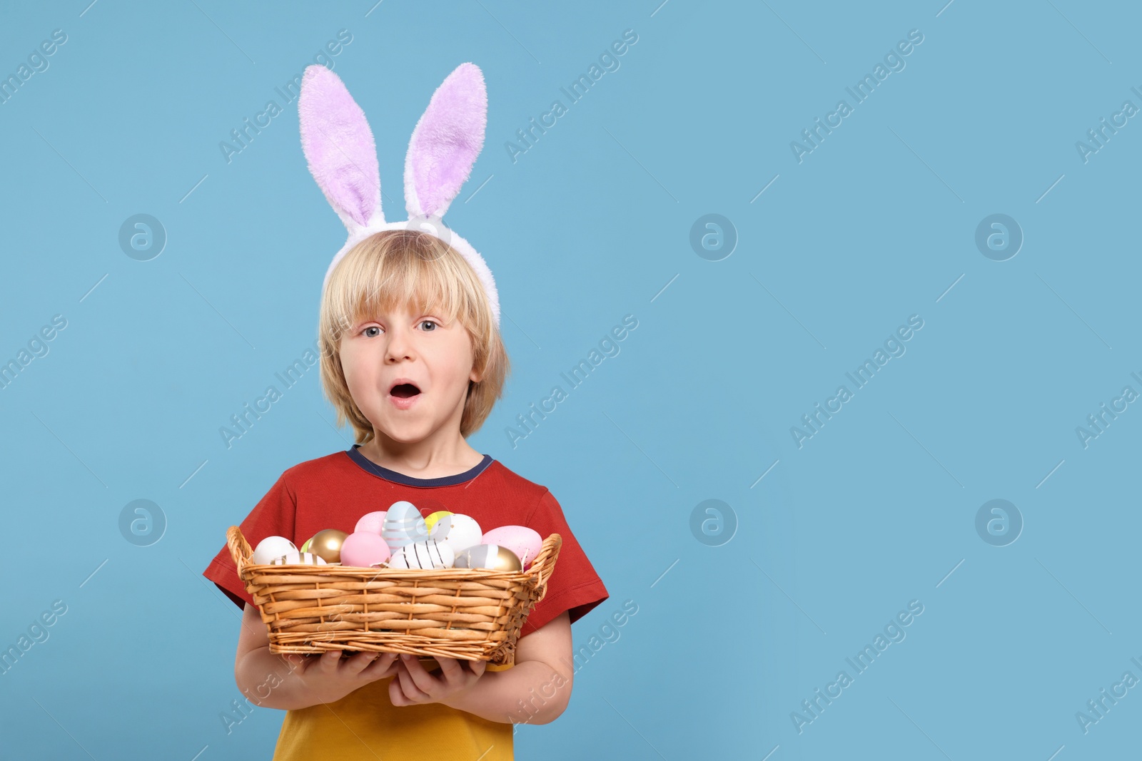 Photo of Happy boy in bunny ears headband holding wicker basket with painted Easter eggs on turquoise background. Space for text