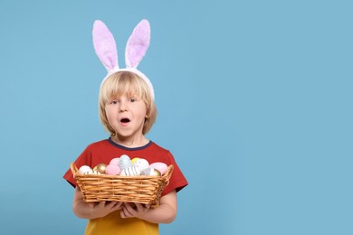 Happy boy in bunny ears headband holding wicker basket with painted Easter eggs on turquoise background. Space for text