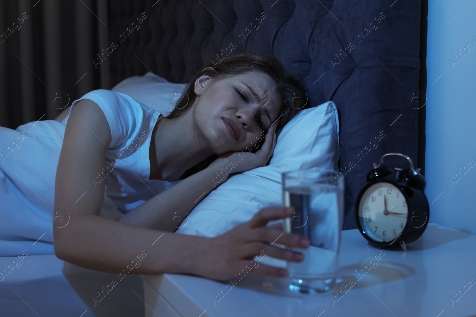 Photo of Young woman with terrible headache at night taking glass of water from stand