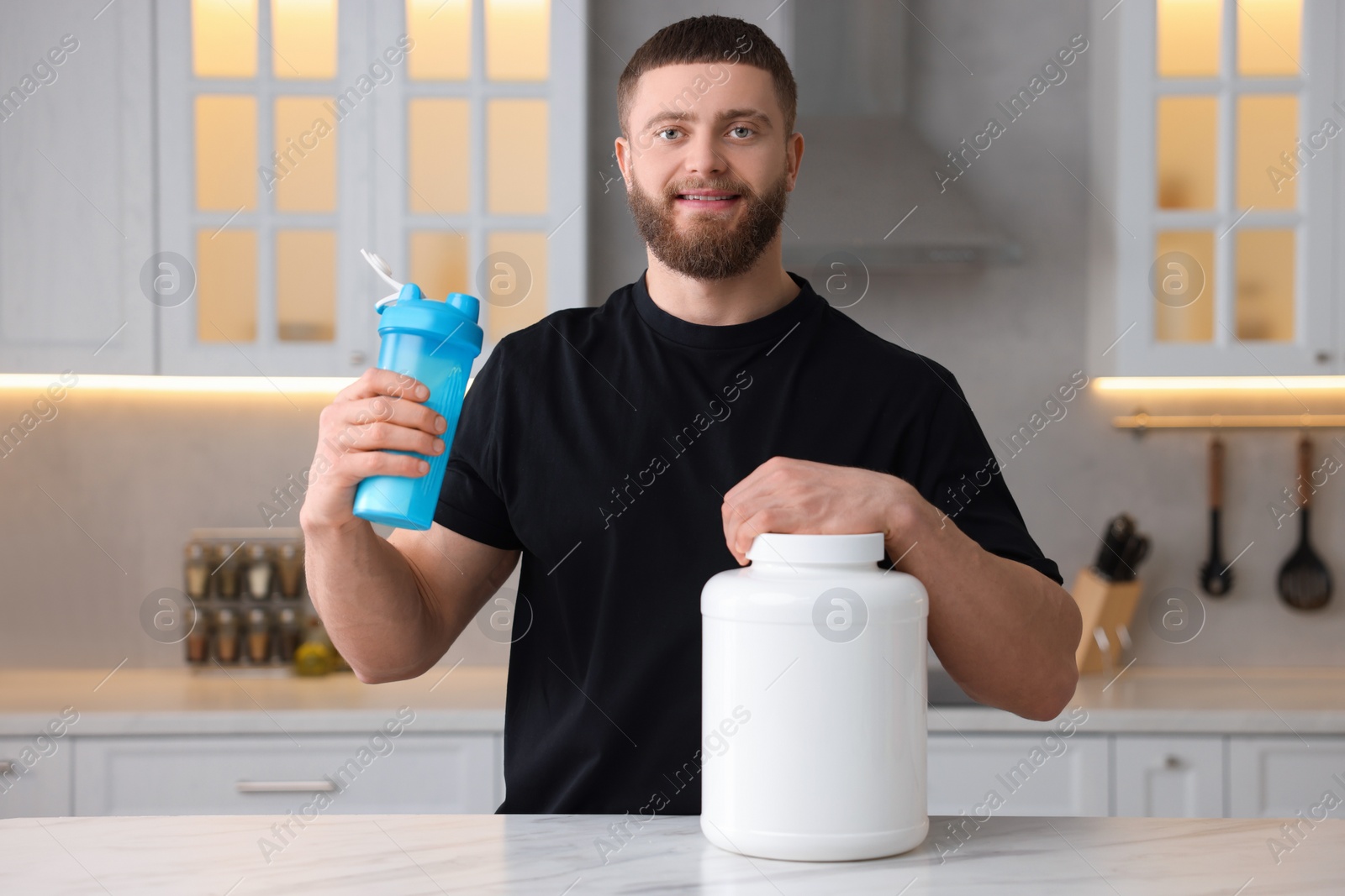 Photo of Young man with shaker of protein and powder at white marble table in kitchen