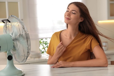 Photo of Woman enjoying air flow from fan at table in kitchen. Summer heat