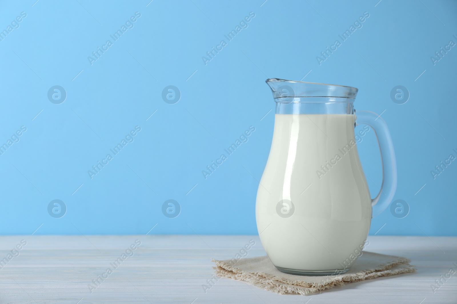 Photo of Jug of fresh milk on white wooden table against light blue background, space for text