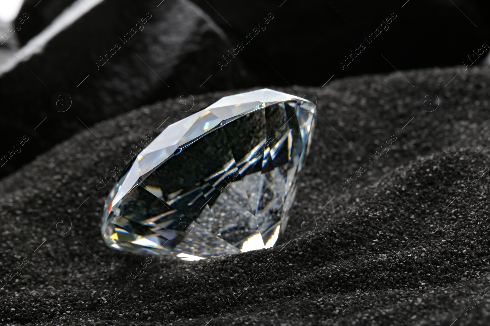 Photo of Beautiful shiny diamond on decorative black sand, closeup