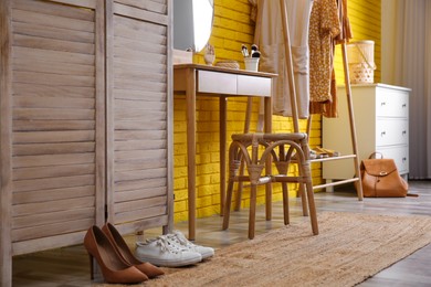 Photo of Wooden stool, dressing table and rack with clothes near yellow brick wall indoors