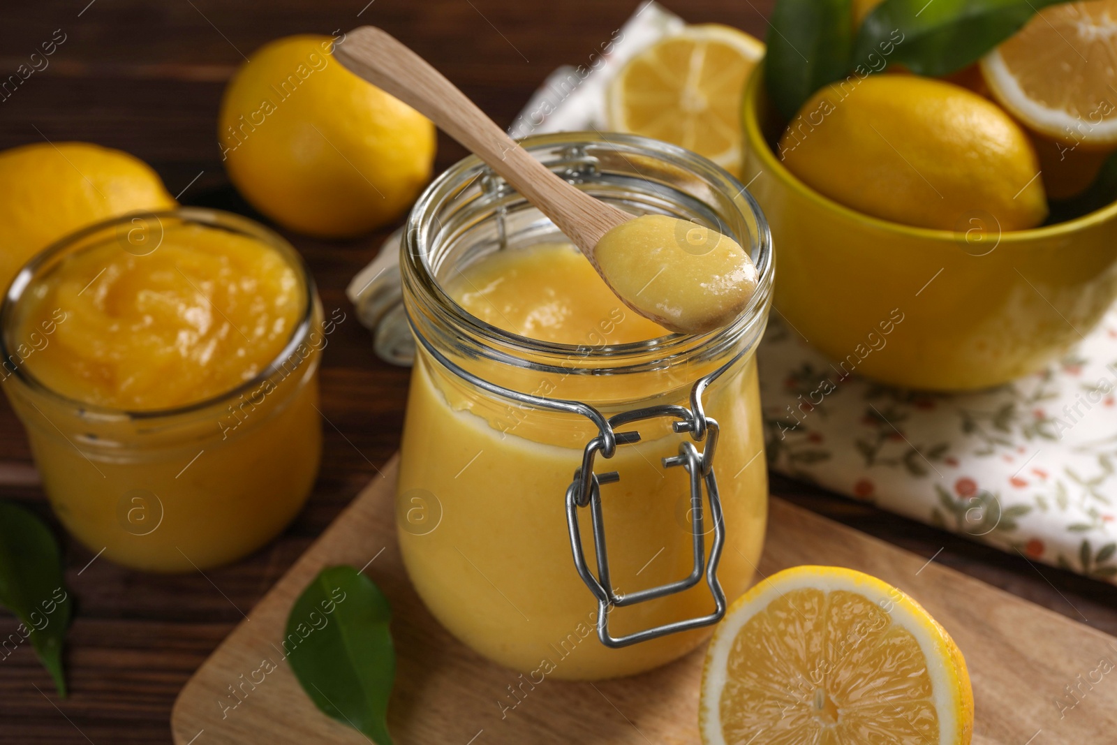 Photo of Delicious lemon curd in glass jars, spoon, fresh citrus fruits and green leaves on wooden table