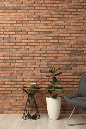 Stylish room interior with beautiful plant, chair and side table near brick wall. Space for text