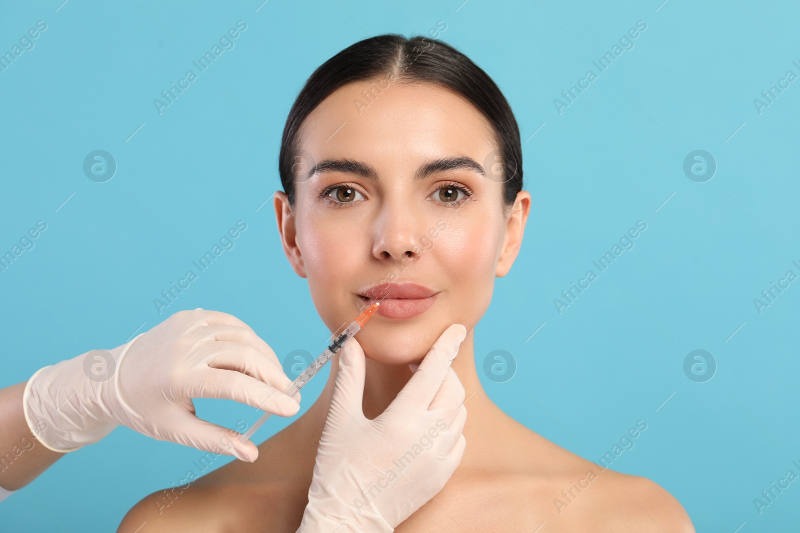 Photo of Doctor giving lips injection to young woman on light blue background. Cosmetic surgery