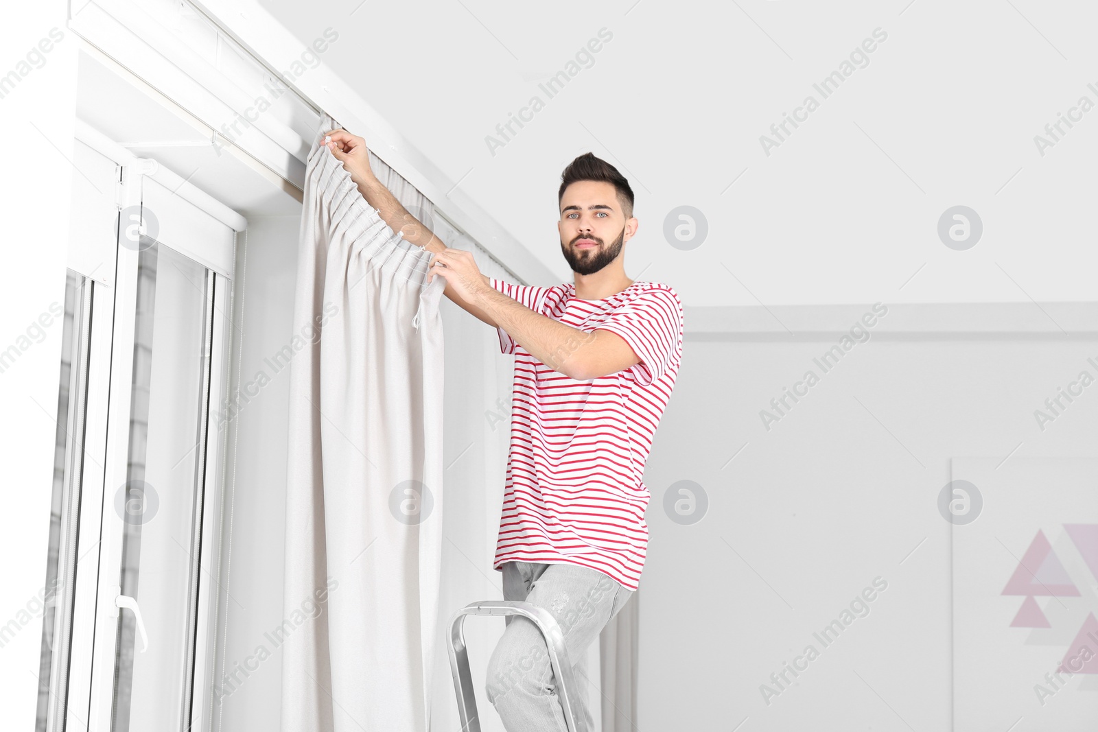 Photo of Young handsome man hanging window curtain in room