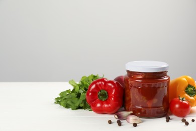 Photo of Glass jar of delicious canned lecho and fresh ingredients on white wooden table. Space for text