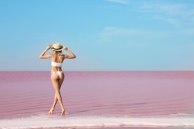 Beautiful woman in swimsuit standing near pink lake on sunny day