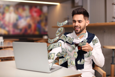 Image of Man with modern laptop and flying dollar banknotes at table indoors. People make money online