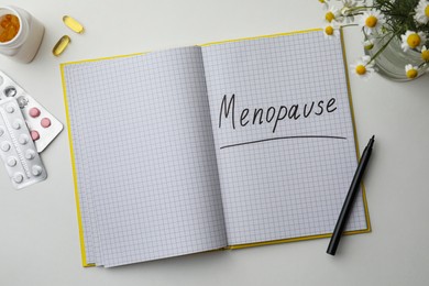 Photo of Notebook with word Menopause, pills, pen and flowers on white background, flat lay
