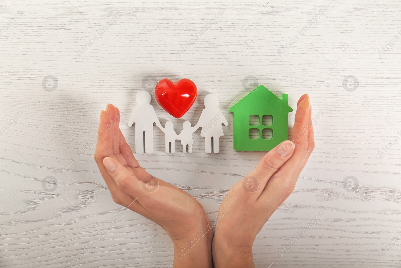 Photo of Woman holding hands near red heart and figures of house and family on white wooden background, top view