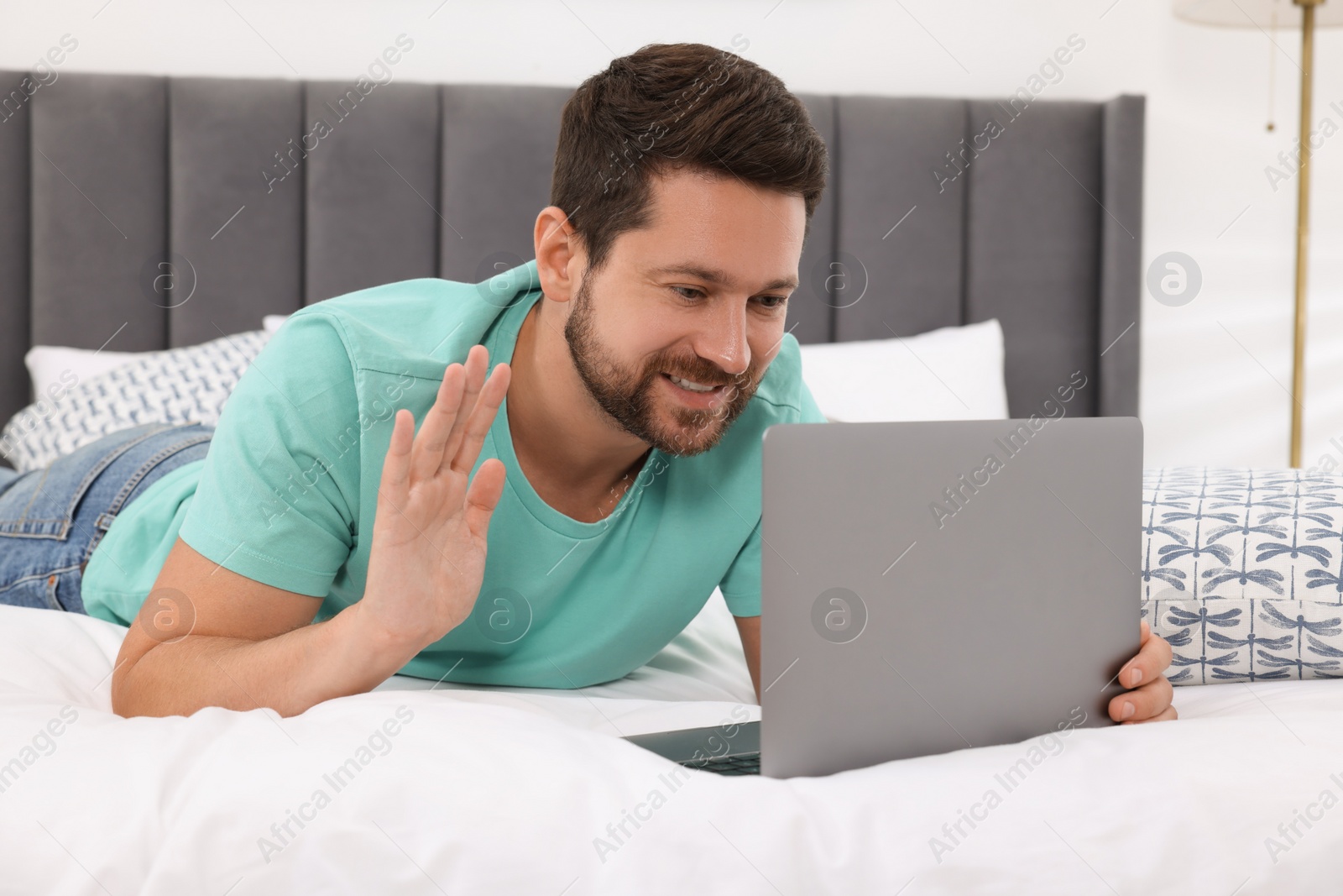 Photo of Happy man greeting someone during video chat via laptop in bedroom