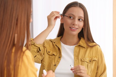 Photo of Beautiful woman applying serum onto eyelashes near mirror indoors