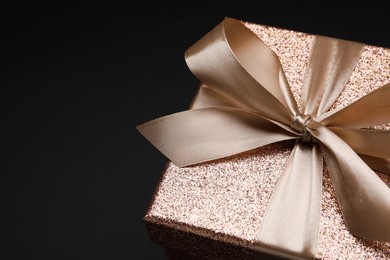 Shiny gift box with golden bow on black background, closeup. Space for text