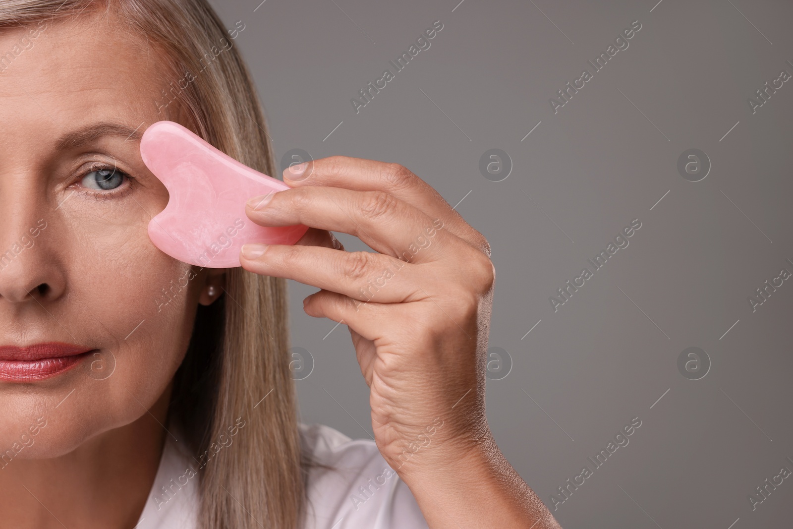 Photo of Woman massaging her face with rose quartz gua sha tool on grey background, closeup. Space for text