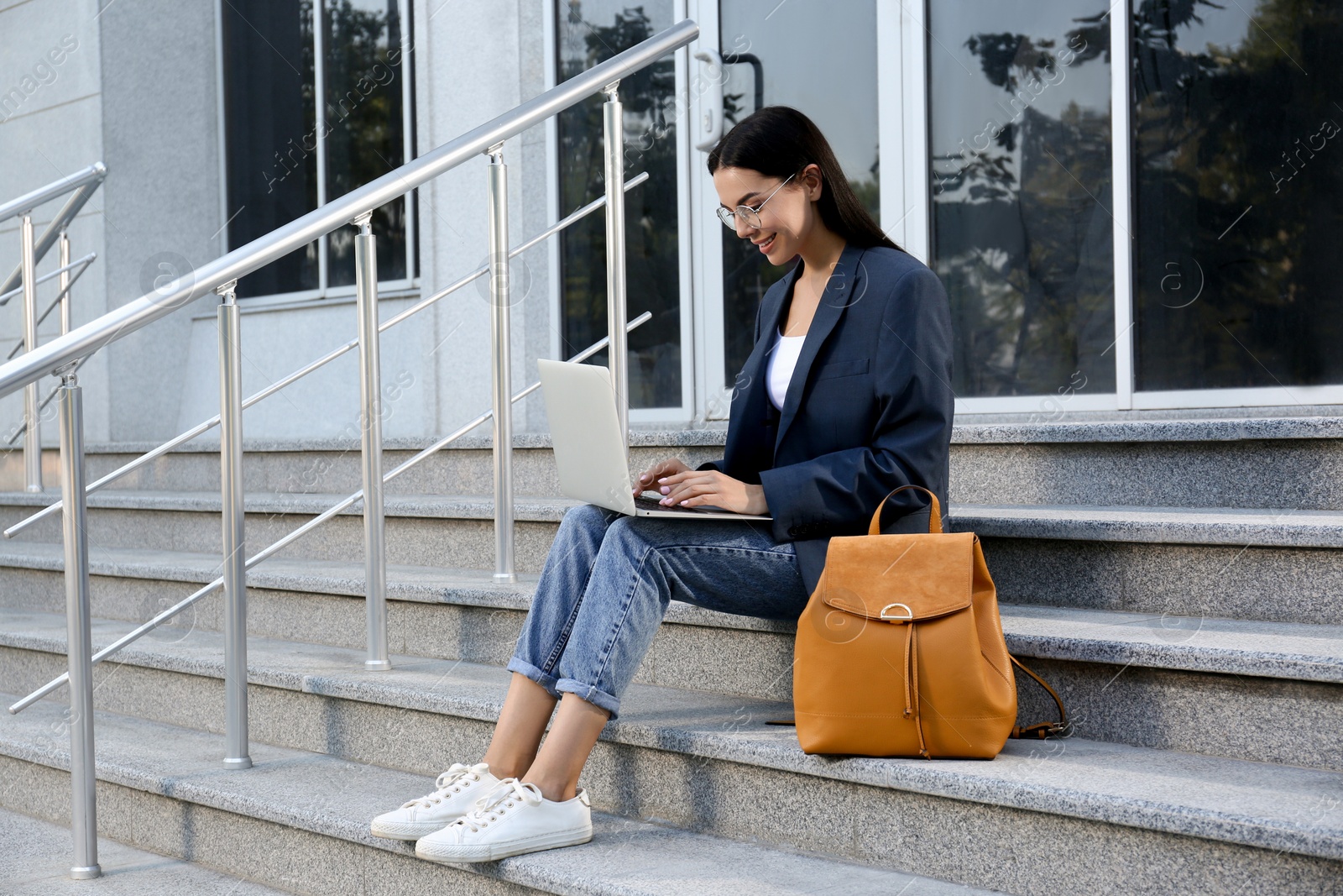 Photo of Beautiful young woman with stylish backpack working on laptop near building outdoors