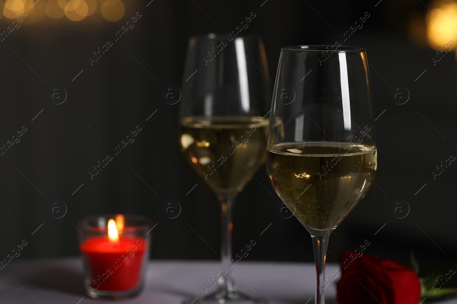 Photo of Glasses of white wine, burning candle and rose flower on table against blurred lights, space for text. Romantic atmosphere