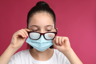 Photo of Little girl wiping foggy glasses caused by wearing medical face mask on pink background. Protective measure during coronavirus pandemic