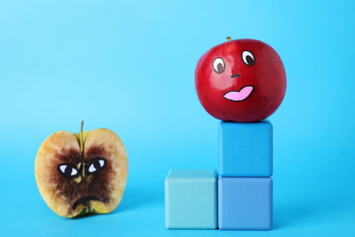 Photo of Ripe and rotten apples with drawn faces on light blue background. Concept of jealousy