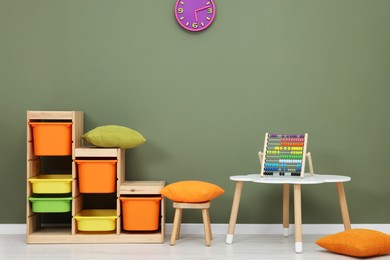 Table with chair, shelves and abacus toy near olive wall in playroom. Stylish kindergarten interior