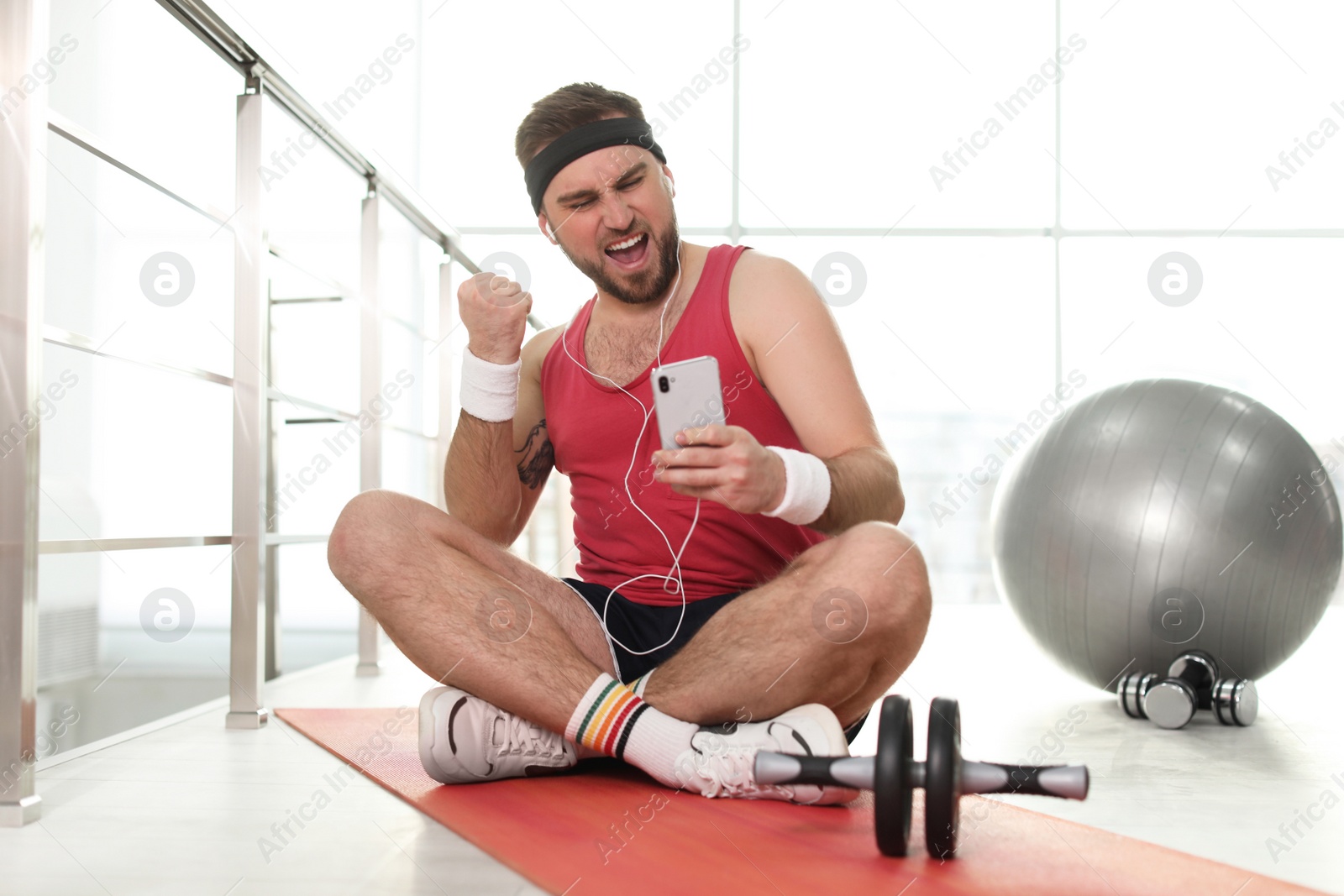 Photo of Lazy young man with sport equipment using smartphone on yoga mat