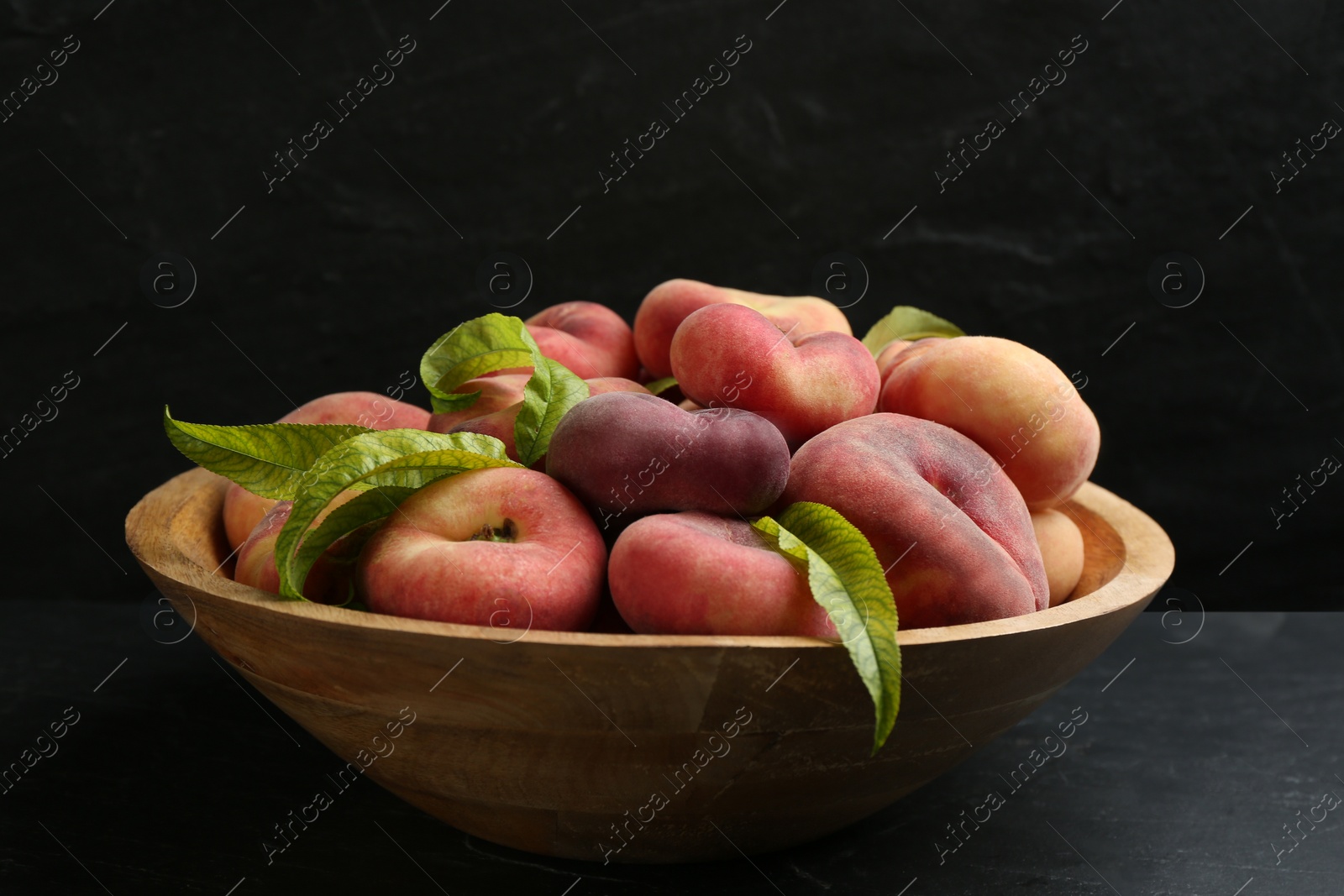 Photo of Fresh ripe donut peaches with leaves in bowl on dark table