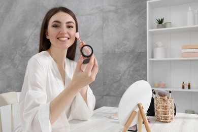 Photo of Beautiful young woman applying blusher with brush at dressing table indoors