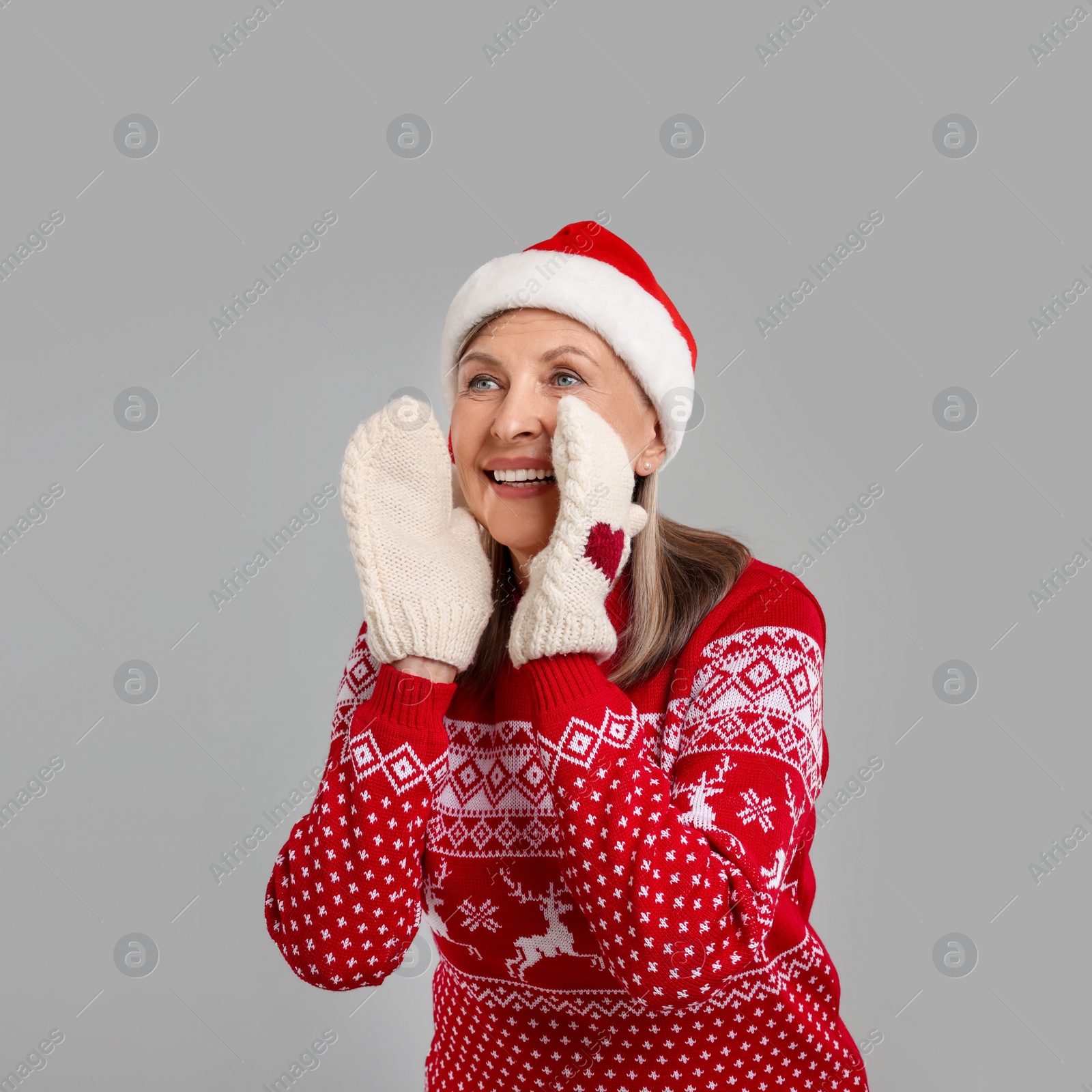 Photo of Happy senior woman in Christmas sweater, Santa hat and knitted mittens shouting something on grey background. Space for text