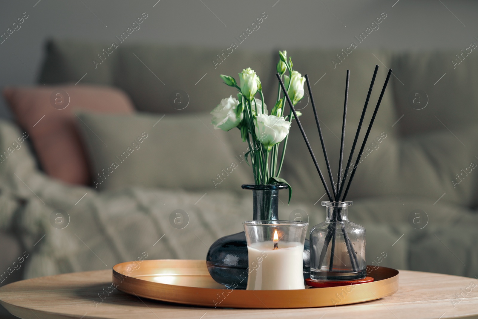Photo of Burning candle with oil reed diffuser and bouquet on wooden table in living room