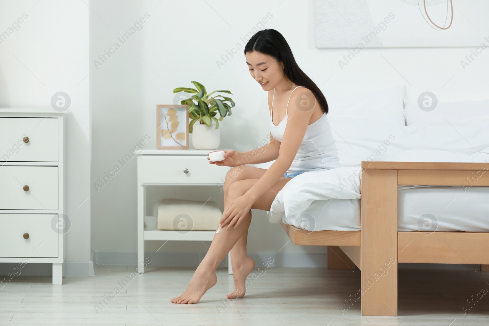 Photo of Beautiful young Asian woman applying body cream on leg in bedroom, space for text