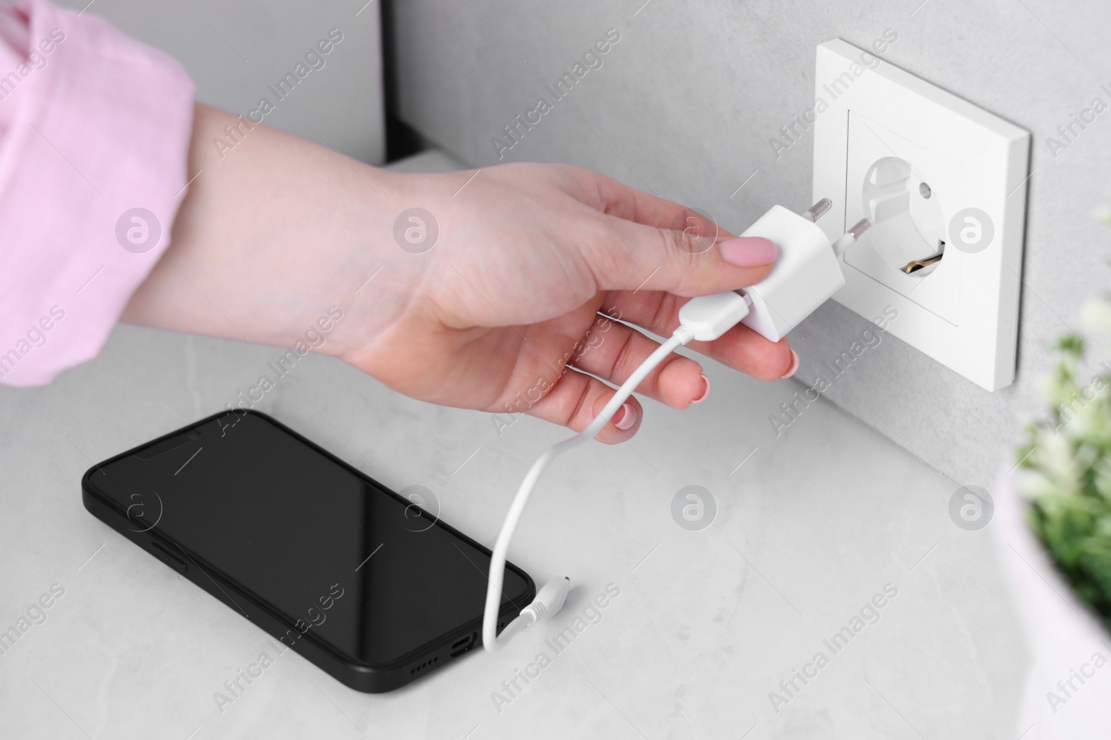 Photo of Woman plugging smartphone into power socket at white table indoors, closeup