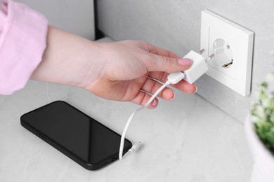 Photo of Woman plugging smartphone into power socket at white table indoors, closeup