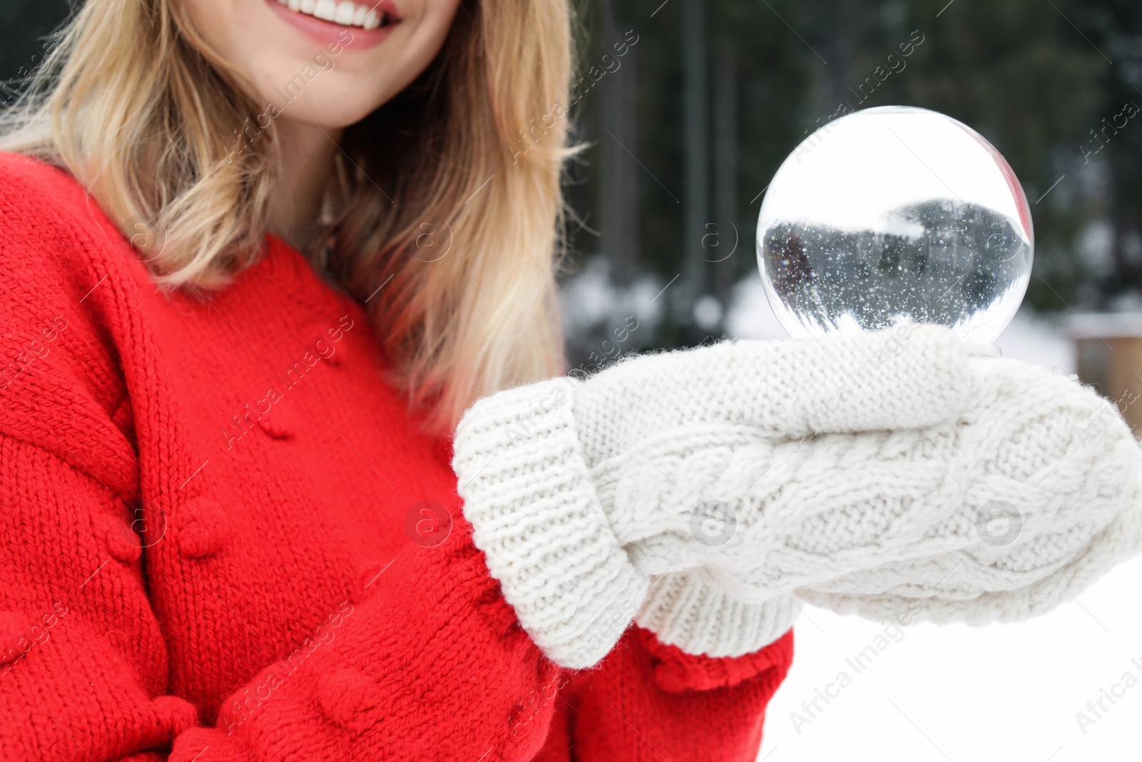 Photo of Woman with knitted mittens holding snow globe outdoors, closeup. Winter vacation