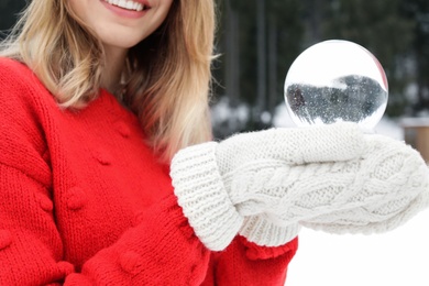 Woman with knitted mittens holding snow globe outdoors, closeup. Winter vacation