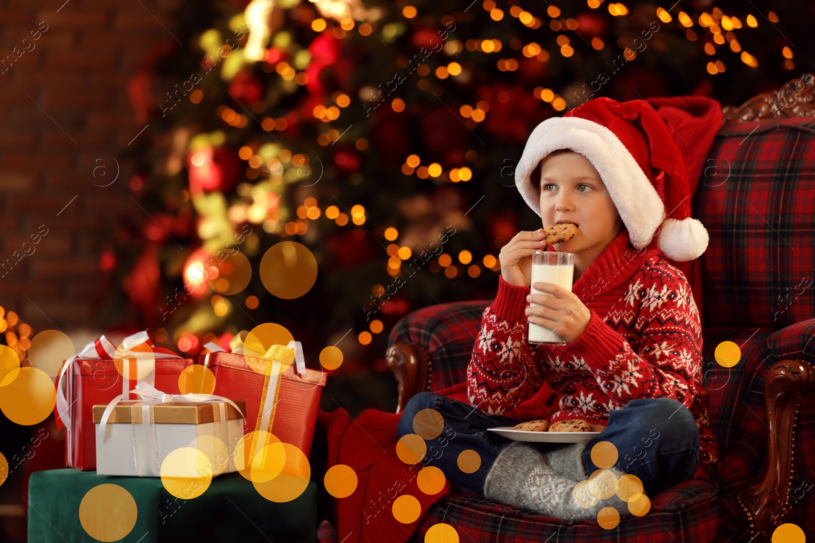 Photo of Little boy in Santa Claus cap with milk and cookies near Christmas gifts at home