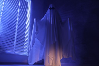 Creepy ghost. Woman covered with sheet near window in blue light, low angle view